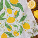 The Gingiber Lemon Grove Tea Towel by Gingiber, made of flour sack cotton and adorned with a vibrant lemon and green leaf pattern, is displayed on a wooden surface beside a halved lemon, whole lemon, and small white flowers.