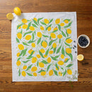 The Gingiber Lemon Grove Tea Towel by Gingiber, adorned with vibrant lemons and green leaves, is displayed on a wooden surface, surrounded by a whole lemon, lemon halves, a bowl of blueberries, and white flowers.