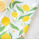 The Gingiber Lemon Grove Tea Towel by Gingiber features bright yellow lemons and green leaves on a light background, with a sliced lemon partially visible in the upper left corner, adding charm to your kitchen décor.