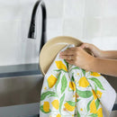 A person dries a beige plate with the Gingiber Lemon Grove Tea Towel by Gingiber, which features yellow lemons and green leaves, next to a black faucet and sink in a modern kitchen.