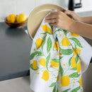 A person dries a beige plate with the Gingiber Lemon Grove Tea Towel by Gingiber, featuring yellow lemons and green leaves. A bowl of lemons rests on a gray countertop in the background.