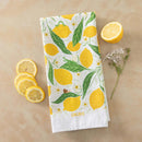 The Gingiber Lemon Grove Tea Towel, made of white flour sack cotton and featuring yellow lemons with green leaves, is neatly folded on a tan surface beside lemon slices, a halved lemon, and white flowers. Brand: Gingiber.