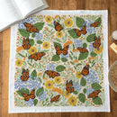 The Gingiber Monarch Garden Tea Towel, made of flour sack cotton, displays orange butterflies with yellow and blue flowers and green leaves—perfect for adding charm to any kitchen décor.