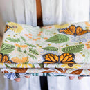 A person in a white shirt holds the Gingiber Monarch Garden Tea Towel, a soft flour sack cotton towel featuring orange monarch butterflies, green leaves, and yellow flowers.