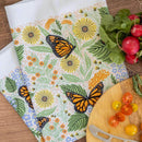 The Gingiber Monarch Garden Tea Towel set by Gingiber features orange and black monarch butterflies, perfect for adding charm to any kitchen décor. Shown with fresh radishes, sliced tomatoes, and a cheese knife on a wooden surface.