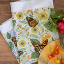 The Gingiber Monarch Garden Tea Towel by Gingiber features yellow flowers, green leaves, and orange butterflies. Shown on wood with radishes and a cutting board, it’s a perfect decorative accent for any kitchen space.