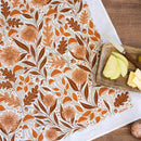 The Gingiber Mushroom Medley Tea Towel by Gingiber, featuring an autumnal botanical motif in brown and orange, lies on a table beside a wooden board with sliced green apples, cheese, and a knife.