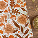 The Gingiber Mushroom Medley Tea Towel by Gingiber, featuring an autumn design with brown and orange leaves, mushrooms, acorns, and flowers, is displayed on a wooden surface next to scattered lentils and a wooden bowl of lentils.