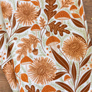 The Gingiber Mushroom Medley Tea Towel by Gingiber features an autumn-inspired design with orange mushrooms, brown acorns, oak leaves, and dandelion-like florals accented by green vines on white—ideal for brightening up your kitchen.