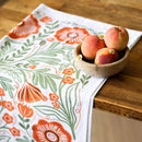 A wooden bowl with three peaches and a small flower rests on a rustic table beside the Gingiber Poppy Tea Towel by Gingiber, made of flour sack cotton and featuring charming orange and green floral patterns.