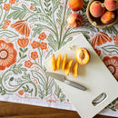 A white cutting board with sliced peach and knife rests on a floral-patterned tablecloth next to whole peaches, small flowers, and the Gingiber Poppy Tea Towel by Gingiber.