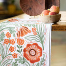 The Gingiber Poppy Tea Towel by Gingiber, made from flour sack cotton and showcasing orange and green floral and butterfly designs, is displayed on a wooden table beside a bowl of peaches and daisies for a charming kitchen accent.