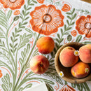 A wooden bowl of peaches and white flowers sits on the Gingiber Poppy Tea Towel by Gingiber, featuring orange flowers and green leaves. Three peaches rest on the tea towel outside the bowl.