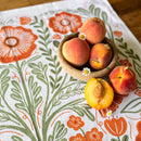 A wooden bowl with four peaches sits on the Gingiber Poppy Tea Towel by Gingiber, made of white flour sack cotton with orange and green floral designs. One peach is halved, surrounded by small white and yellow daisies.