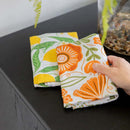 A hand picks up the Gingiber Poppy Tea Towel, a colorful flour sack cotton towel with an orange floral pattern, from a dark table beside another kitchen towel with lemons. A glass vase with plants and pebbles sits nearby. Brand: Gingiber.