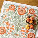 A wooden table displays a small brown bowl with peaches and peach blossoms, set atop a white Gingiber Poppy Tea Towel by Gingiber featuring orange and green floral patterns.