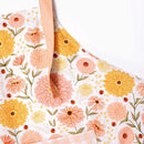 A close-up of the Gingiber Zinnia Apron by Gingiber, made from soft cotton and featuring a yellow, orange, and pink floral pattern, with light pink adjustable straps and a pink-and-white gingham section at the bottom.