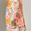 A close-up of the Gingiber Zinnia Apron by Gingiber, a cotton cooking apron with a yellow, peach, and pink floral pattern featuring large flowers and ladybugs. A pink flower is tucked into its adjustable checkered pocket.