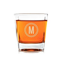 A square Whiskey Glasses: Medalist by Swanky Badger filled with amber liquid, etched with a circular emblem featuring the letter M in the center.