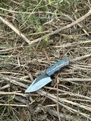 The Texan Knives Good Hunting Knives features a 9" handmade J2 steel blade and a blue-orange epoxy resin handle, shown resting on dry grass with its leather sheath.