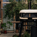 The Gozney Dome XL Gen 2 Door by Gozney, featuring a chimney and insulated oven door on a black stand, sits by potted plants and a wooden deck—ideal for smoking and slow-roasting outdoors.