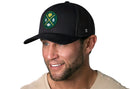 A smiling man with a short beard wears a Haka Hat Green Bay Trucker Hat—a black mesh snapback featuring a green and yellow "GB" Cheese X Football patch. He is also wearing a light tan shirt.