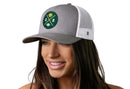 A woman with long brown hair smiles while wearing the Haka Hat Green Bay Trucker Hat, a gray and white snapback featuring a round green patch with yellow and white GB Cheese X Football symbols on the front.