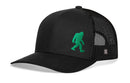 The Haka Hat Green Bigfoot Trucker Hat is a black mesh snapback with a curved brim, featuring a green embroidered Sasquatch silhouette on the front left panel.