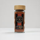 A glass jar of Spice Wheel Guajillo Powder from the brand Spice Wheel, crafted using dried guajillo chilies for a mild to medium heat, rests on a white surface. The jar is topped with a wooden lid and displays a black and red label featuring a small chili icon and the net weight of 55g.