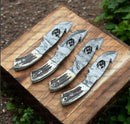 Four Texan Knives, including the Custom Handmade Guthook Hunter Knife with Damascus steel blade and stag horn handle, are displayed side by side on a wooden surface outdoors, set against green foliage.