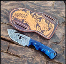The Texan Knives Premium 9" Damascus Guthook Skinning Knife with a blue resin handle lies next to its brown leather, floral-tooled sheath, displayed on a textured stone surface.