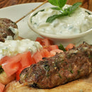 A grilled kebab skewer rests on pita bread with chopped tomatoes, a dollop of tzatziki, and a sprinkle of SALTOPIA Infused Sea Salts Saltopia Za'atar Spice Blend. In the background is a bowl of tzatziki garnished with mint.