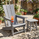 A gray Adirondack Chair with Cup Holder - HDPE Outdoor Seating by DirectDeals holds a pink drink on the armrest, next to a matching table on a stone patio surrounded by potted plants and blooming flowers.