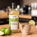 A bottle of Hella Cocktail Co. Habanero Margarita Mix sits next to a salt-rimmed glass of ice garnished with a lime wheel and two whole limes on a wooden surface.