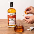 A bottle of Hella Cocktail Co. Old Fashioned Mix sits beside a glass of whiskey with ice. A hand holds an orange peel and a lit match, ready to garnish the drink on a wooden table.