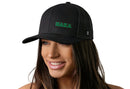 A woman with long brown hair wears a Haka Hat Haka Branded Trucker Hat—Black Rubber Patch Snapback. “HAKA” appears in green on the front. She smiles, looking to the side, and the comfort-fit sweatband ensures all-day ease. The background is white.