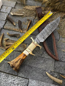 The Texan Knives Custom Handmade Damascus Steel Bowie Knife features a 13-inch patterned blade, bone and brass guard handle, and comes with a leather sheath, shown resting beside measuring tape and antlers on stone tiles.