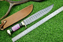 The Texan Knives Damascus Steel "The Bowie" Knife, 16" with brass guard and resin pakkawood handle, lies beside its brown leather sheath and a metal ruler on grass, with an antler in the background.