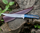 The Texan Knives Custom Handmade Damascus Fillet Knife – 13” with a pine cone handle and leather sheath sits on a weathered tree stump amid green plants.