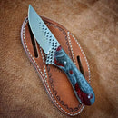 The Texan Knives Handcrafted Cowboy Knife rests on a Fall & Winter Edition leather belt loop sheath by Texan Knives, featuring decorative stitching—ideal for ranch work and displayed on a suede surface.