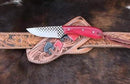 The Texan Knives Handcrafted Cowboy Knife with rasp blade and red resin handle rests on its floral and geometric leather sheath, displayed on a leather belt atop an animal hide. All items are by Texan Knives.