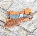 The Texan Knives Ranch Cowboy Knives – Handmade Rasp Tool Blade with Pine Cone Handle 8 rests on a textured brown leather sheath atop a stone surface.