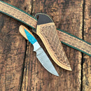 The Texan Knives Handcrafted Back of Belt Knife features a patterned Damascus steel blade and a resin and olive wood handle, displayed on a tooled leather sheath and belt with geometric designs atop a rustic wooden surface.