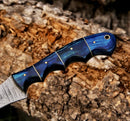 Close-up of the Texan Knives Handmade 8" Rasp Steel Sheep Horn Hunting Knife, featuring a contoured blue handle with brass pins and finger grooves against tree bark. Comes with a leather belt sheath—ideal for outdoor adventures.