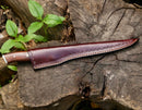 A Premium Custom Handmade Damascus Steel Fillet Knife – 13” Rosewood Handle by Texan Knives, complete with a brown leather sheath, rests on a weathered stump among green leaves—an essential for any outdoor enthusiast.