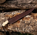 The Texan Knives Fishing Fillet & Boning Knife features a 13" Damascus steel blade, a camel bone and rosewood handle, and comes with a leather sheath. It is displayed on tree bark with stacked logs in the background.