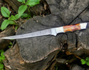 The Texan Knives Custom Handmade Damascus Filleting Knife – 13" features a patterned blade and a polished camel bone handle, shown resting on a weathered tree stump amid green plants. Includes leather sheath.