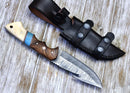The Texan Knives Premium Damascus Guthook Skinning Knife, featuring a 10” patterned blade and a rosewood & bone handle, rests next to its black leather sheath on a weathered wooden surface.