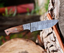 The Texan Knives Custom Handmade 5-Inch Damascus Skinning Knife with a rosewood handle rests on a cut log outdoors, its patterned blade and elegant design showcased against a blurred background.