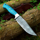 The Texan Knives Custom Handmade Damascus Skinning Knife – 4.5" blade, featuring a patterned steel blade and blue textured handle, rests on tree bark with its leather sheath partially visible amid green leaves and grass.